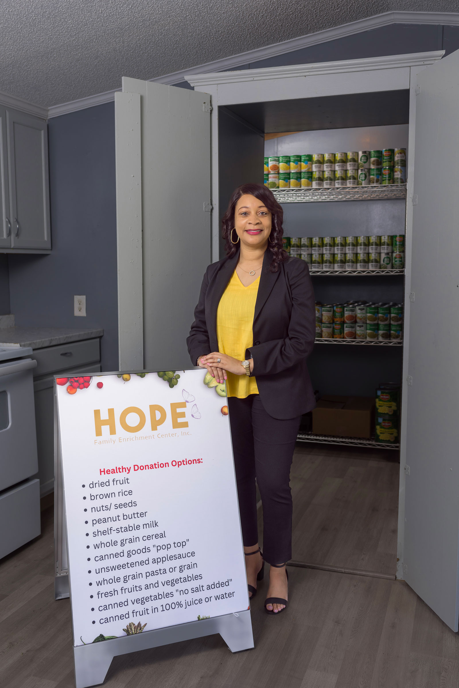 A professionally dressed woman standing in front of many rows of food cans and beside a sign that lists “HOPE.”
