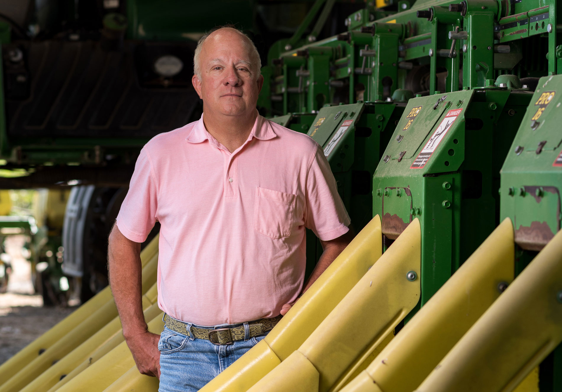 A bald man wearing a pink polo and jeans standing beside farm equipment.