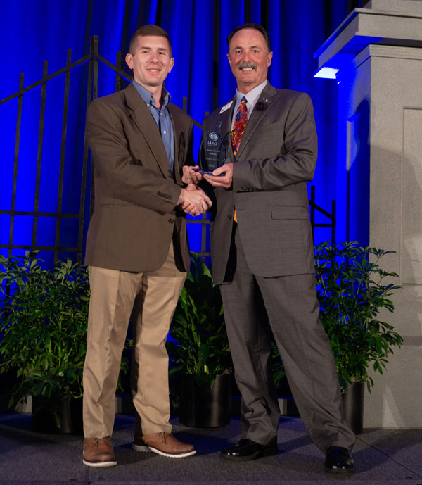 Two standing men smiling and the one on the right holding an award.