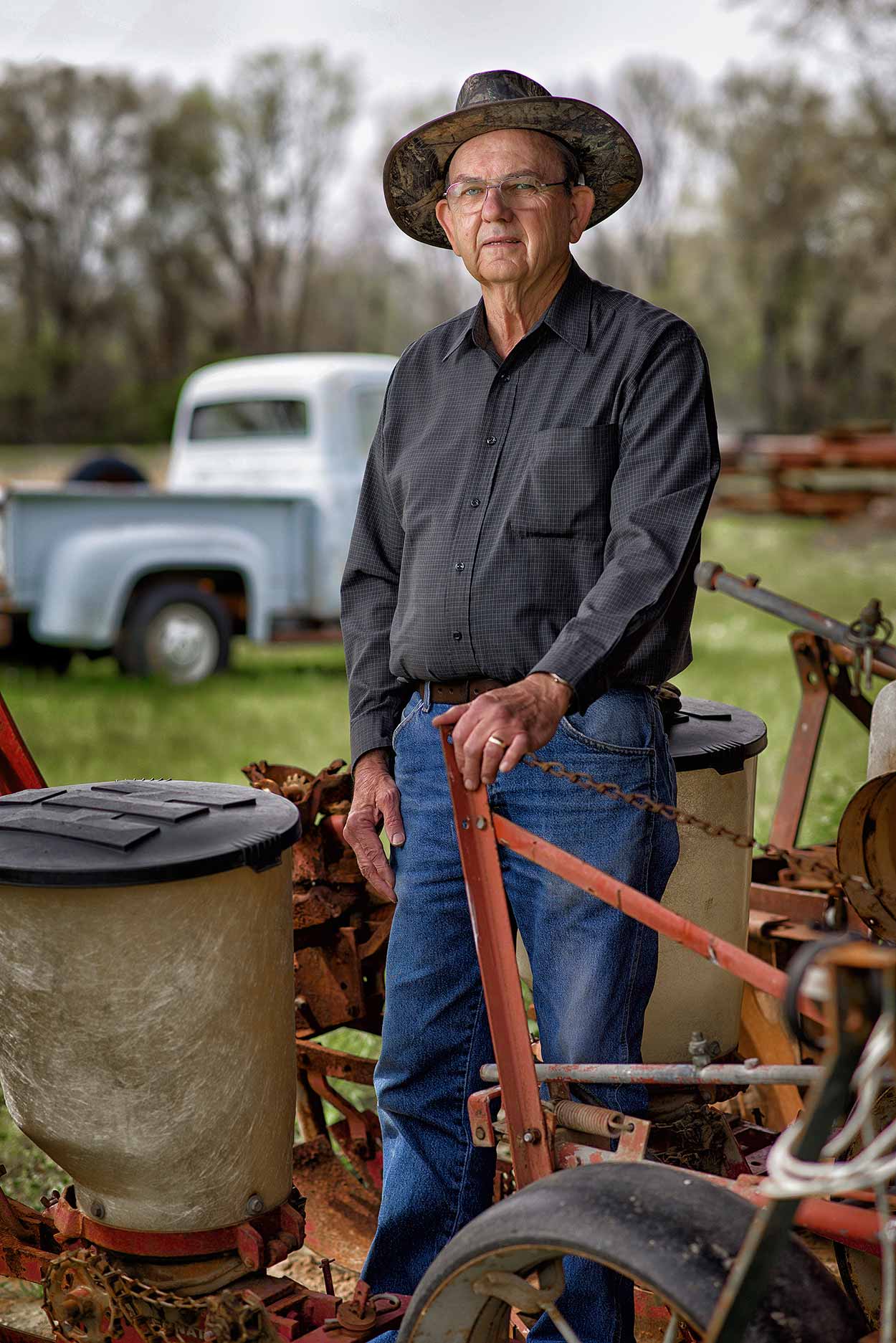 A man in a black shirt and blue jeans wearing a camouflage hat stands in front of an old, light blue truck and rests his hand on an old, rusty tractor. 