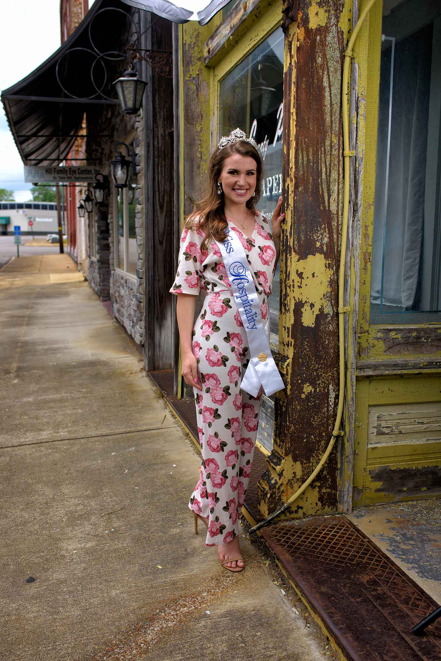 A young woman with brown hair and a silver crown wears a white jumpsuit with pink roses and a white sash embroidered with the words “Miss Hospitality.” She stands with her hand resting on a yellow, chipped wooden storefront.