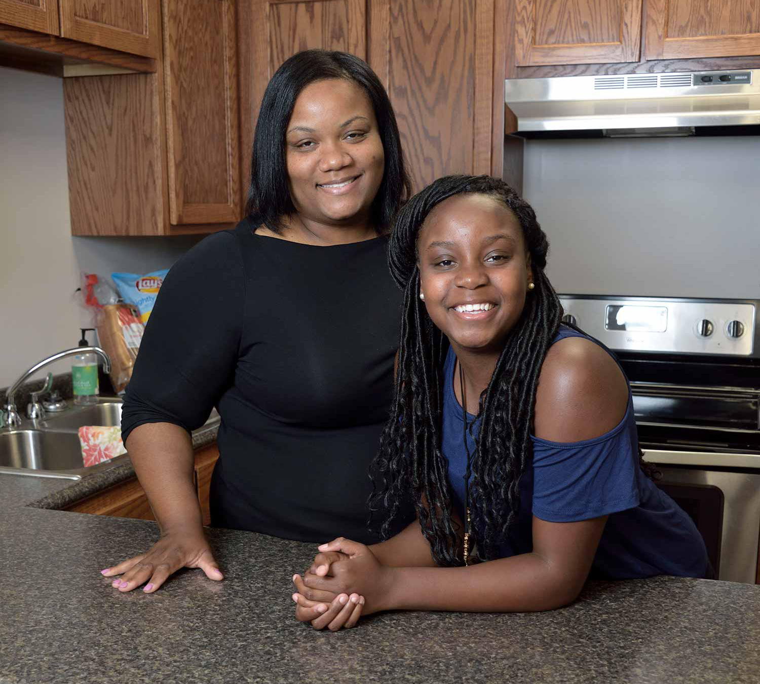 Two females stand behind a kitchen counter. On the left, a woman in a black shirt stands, and on the right, a teen girl wears a blue shirt and leans forward with her arms propped on the counter. 