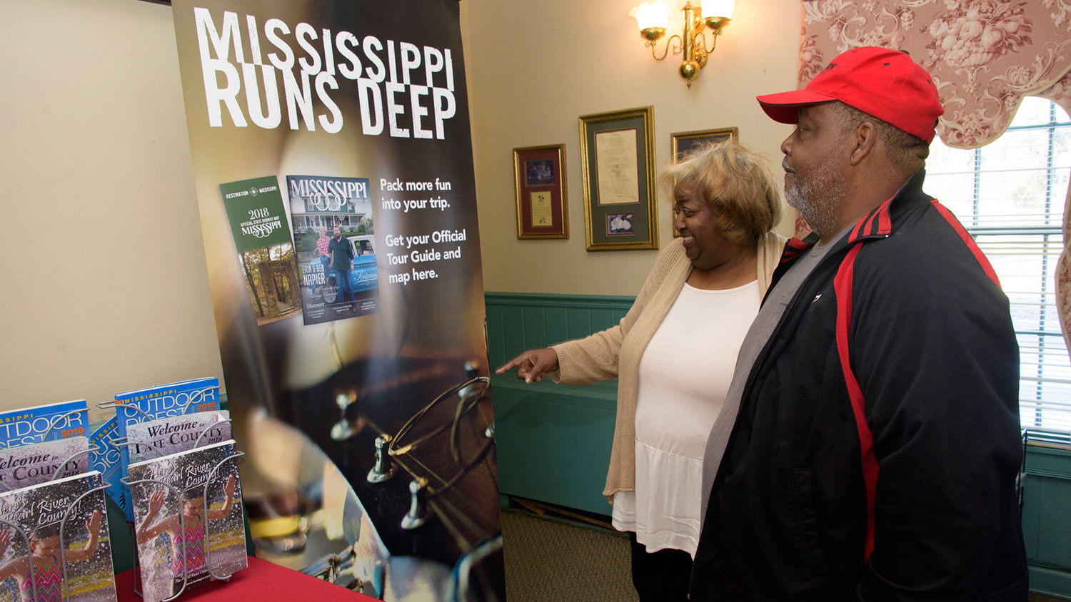 A man wearing a red hat stands next to a smiling woman pointing at a poster that says "Mississippi Runs Deep." 
