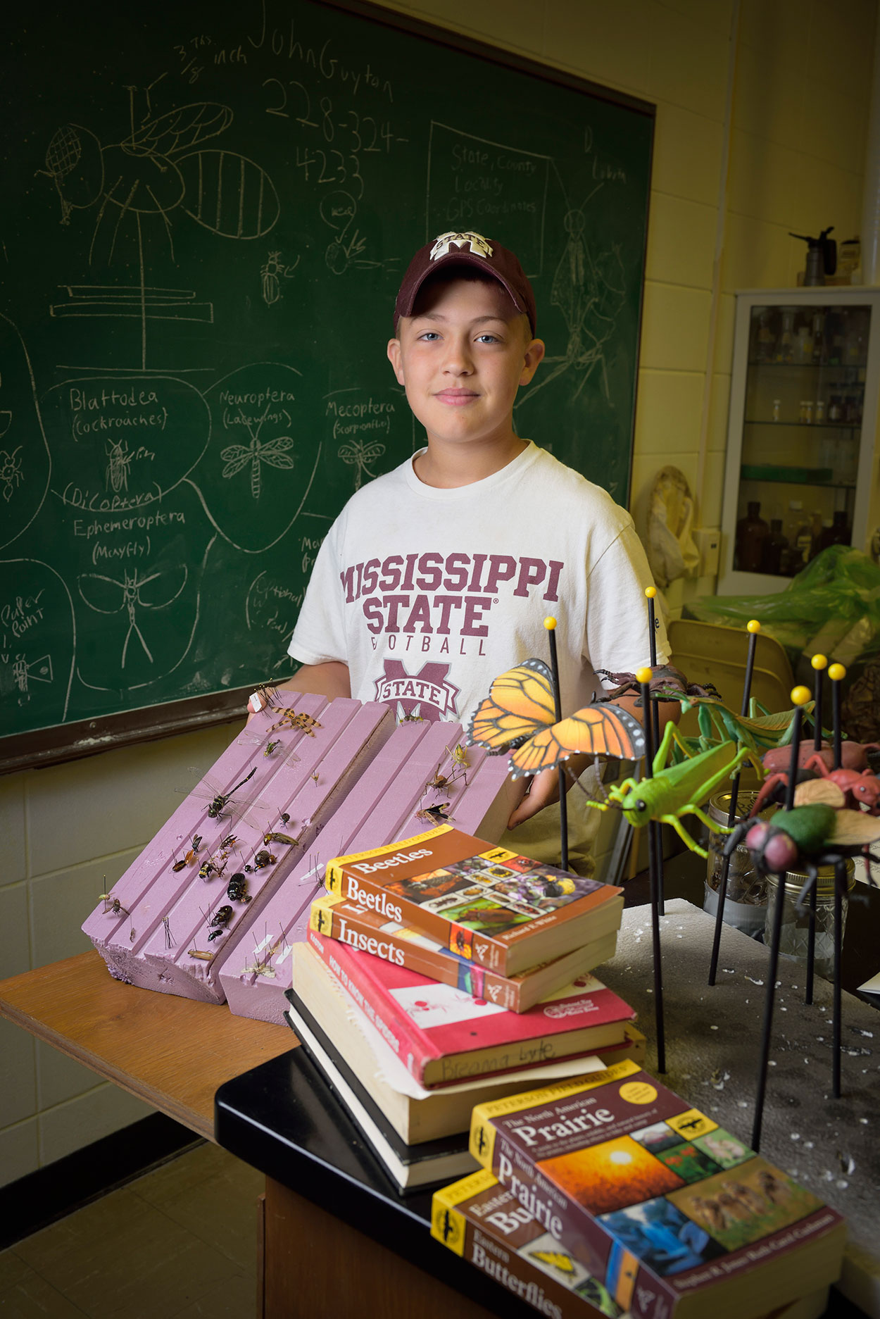 A young boy wearing a Mississippi State hat and t-shirt stands behind a stack of books and insect replicas while he holds his project, a purple piece of wood that showcases many different types of insects.