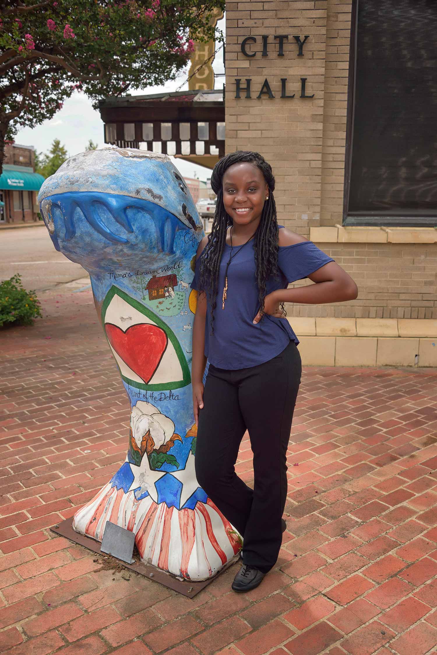 A teen girl with a blue shirt and black pants stands in front of a building labeled “City Hall” and to the right of a statue of a catfish, blue on top with red and white strips on the bottom fin. 