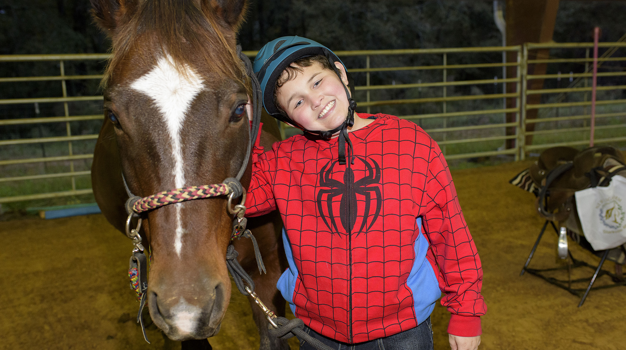 A young boy wearing a red Spiderman jacket and a blue helmet stands next to a brown and white horse.