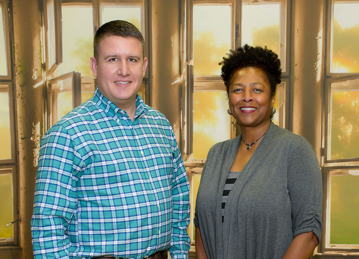 A man wearing a light blue, plaid shirt and a woman wearing a grey shirt stand next to each other, smiling. 