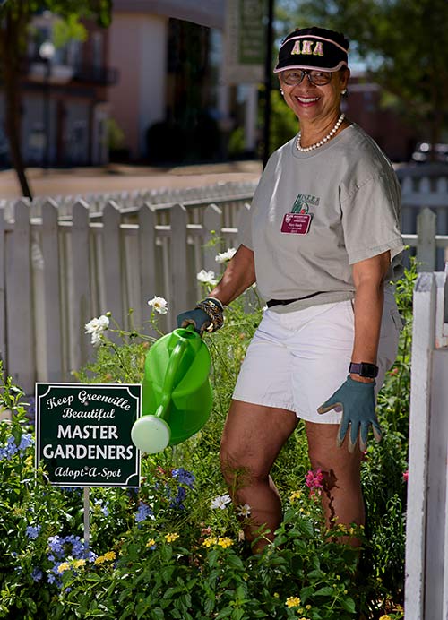 A woman stands next to a flower bed holding a green watering can.