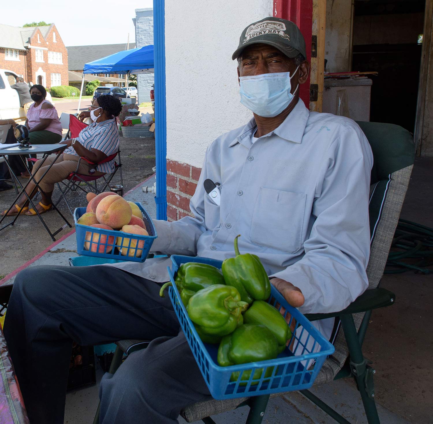 A man wearing a mask holds two blue baskets filled with green bell peppers and peaches.
