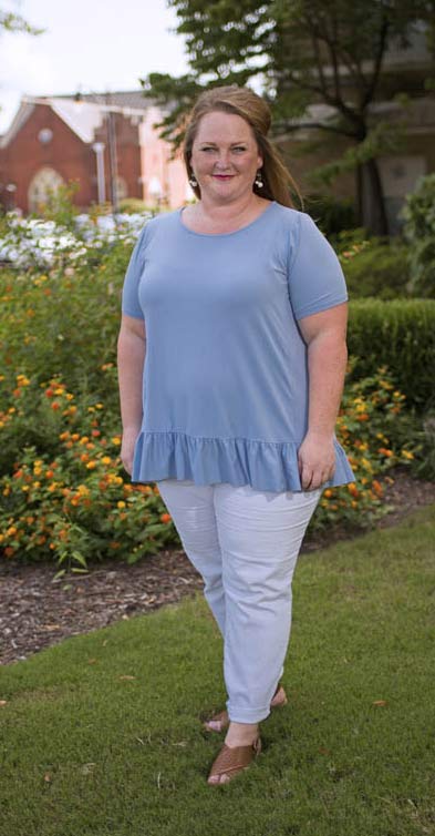 A woman wearing a light blue shirt and white pants stands in green grass in front of flowery bushes.