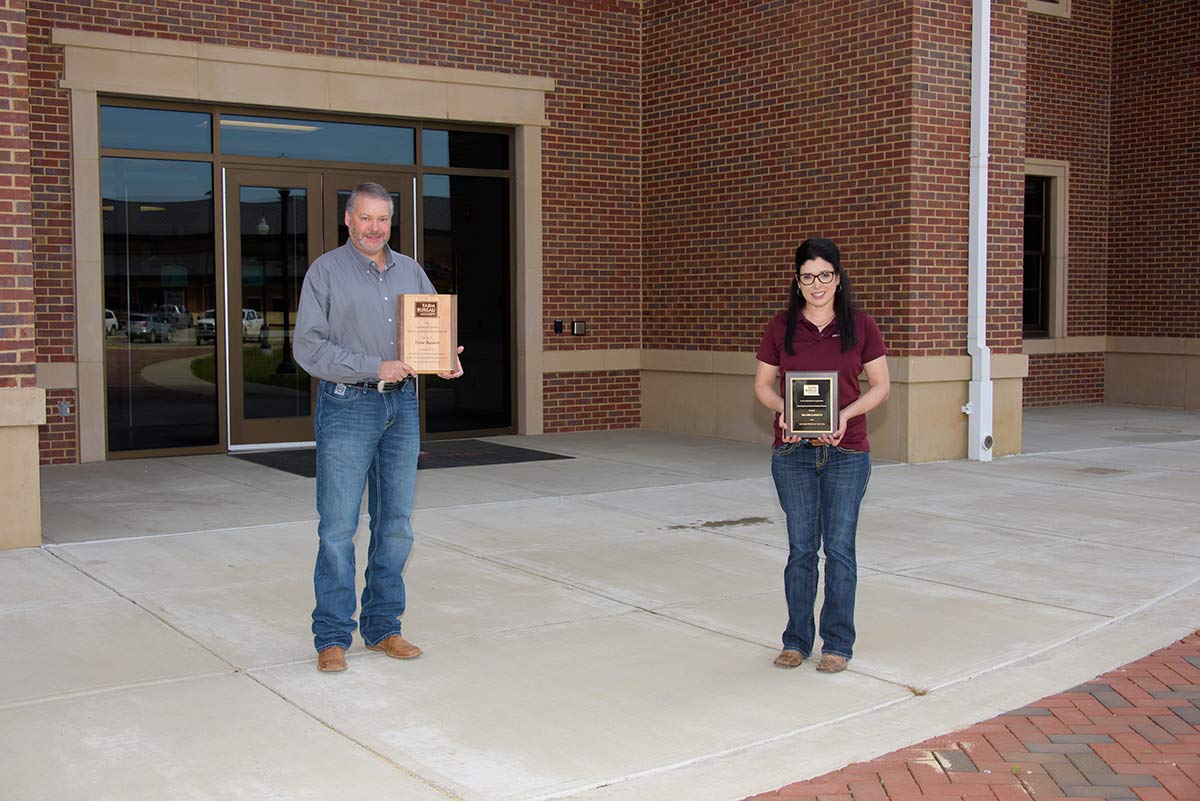 A smiling man and a woman holding plaques stand several feet apart in front of a brick building.