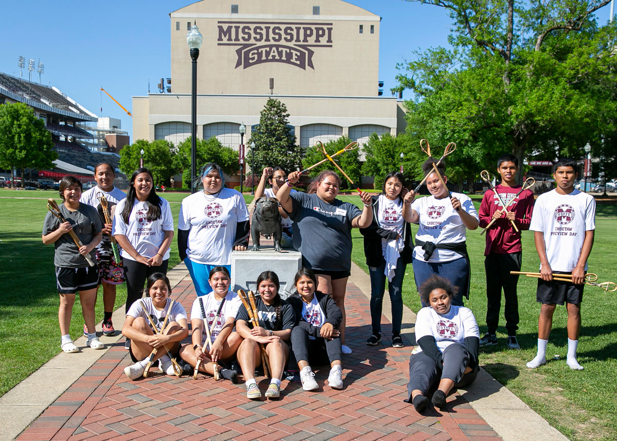 A group of high school students poses for a photo around a bulldog statue in front of the Mississippi State University football stadium. 