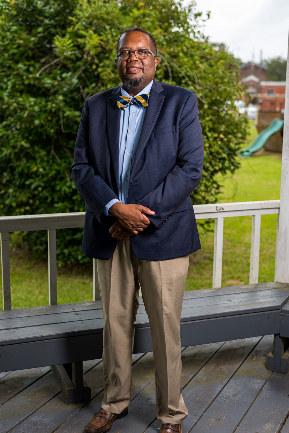 A Black man with glasses wearing a blue and yellow striped tie, a blue dress jacket, and khakis, standing on a deck in front of a tree.