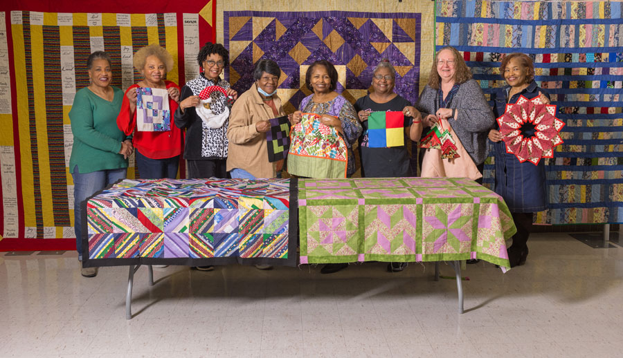 Eight women smiling and standing behind a table with quilts laid on it.