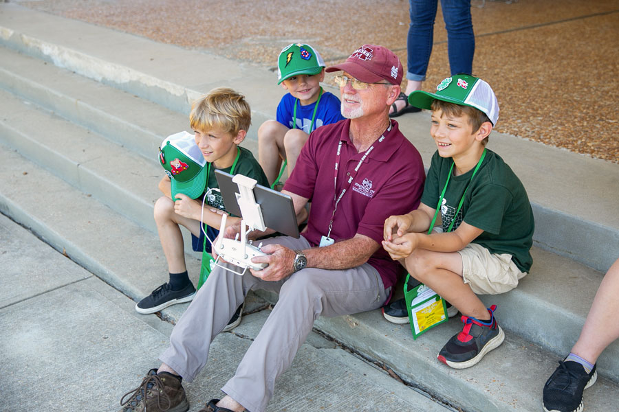 Three boys seated around a man holding a remote for an uncrewed aerial vehicle