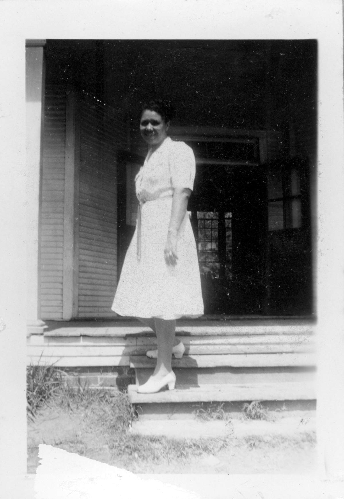An old black and white photograph of a young black woman standing on the stairs of a front porch.