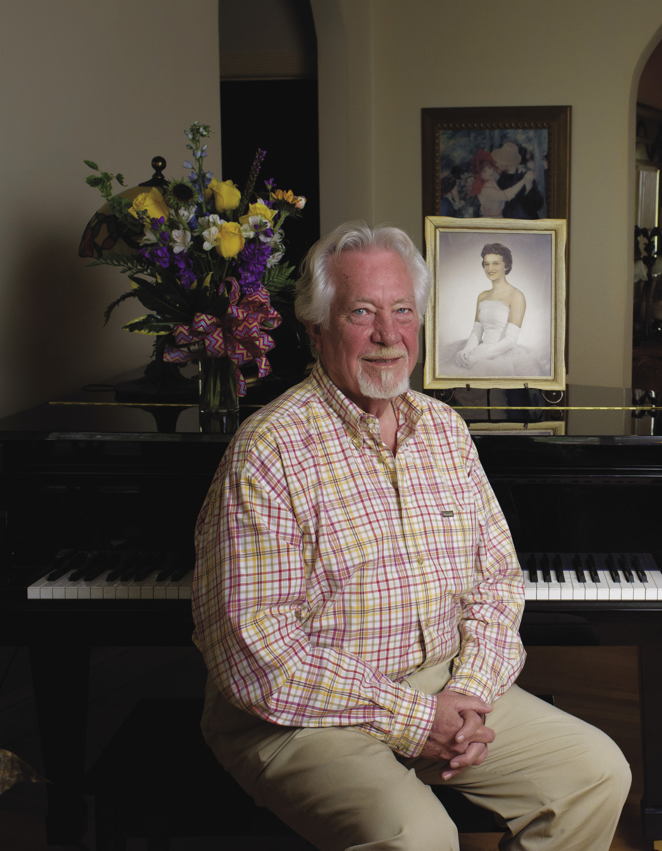 Michael Hale sitting in front of a piano with flowers and a framed picture of late wife, Vicki M. Smith, on top.