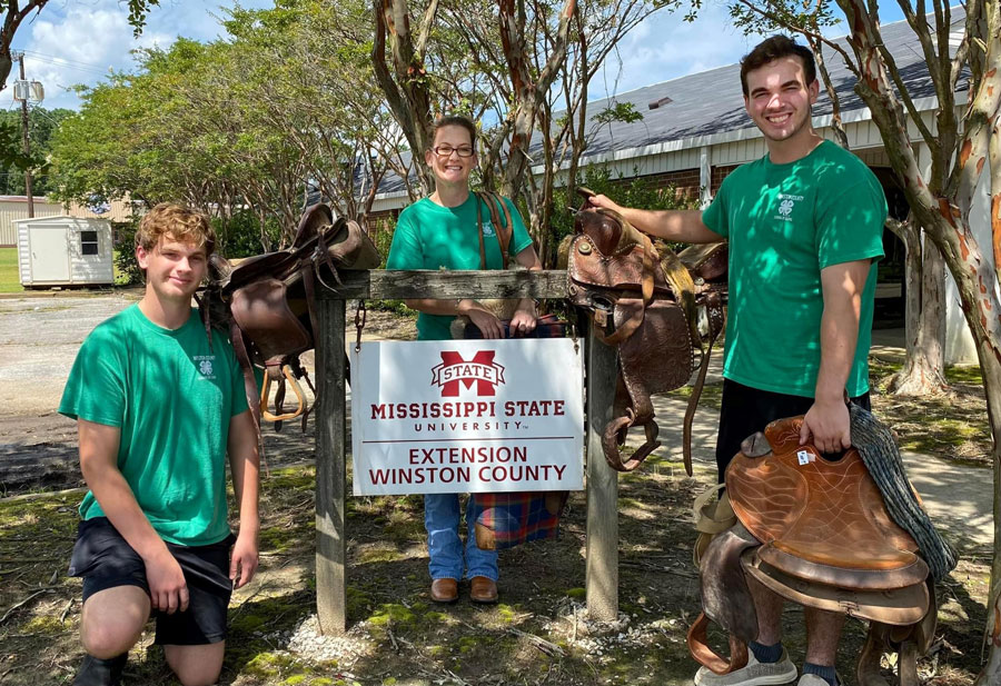 Two boys and a woman, all wearing green shirts, standing with saddles near a Mississippi State University Extension Service sign.