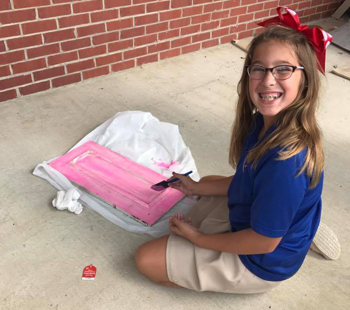 A young girl sits on the ground while painting a piece of wood pink for her deer antler jewelry holder.