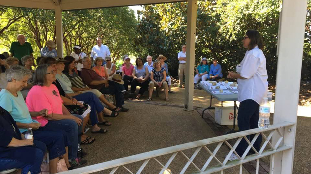 A large group of adults sit under a pavilion and listen as an Extension specialists talks about gardening during an outdoor event 