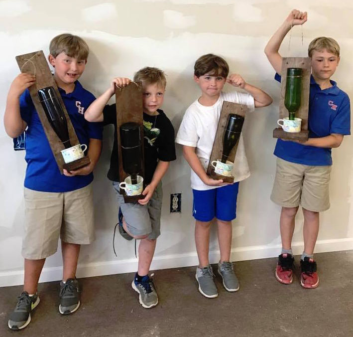 Four young boys stand against a wall while holding bird feeders made out of recyclables.