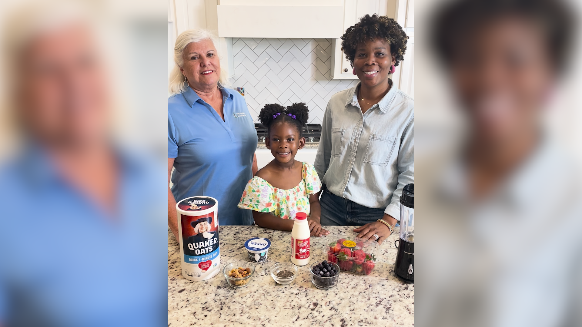 Two women and a child stand behind a kitchen counter filled with ingredients to make a smoothie