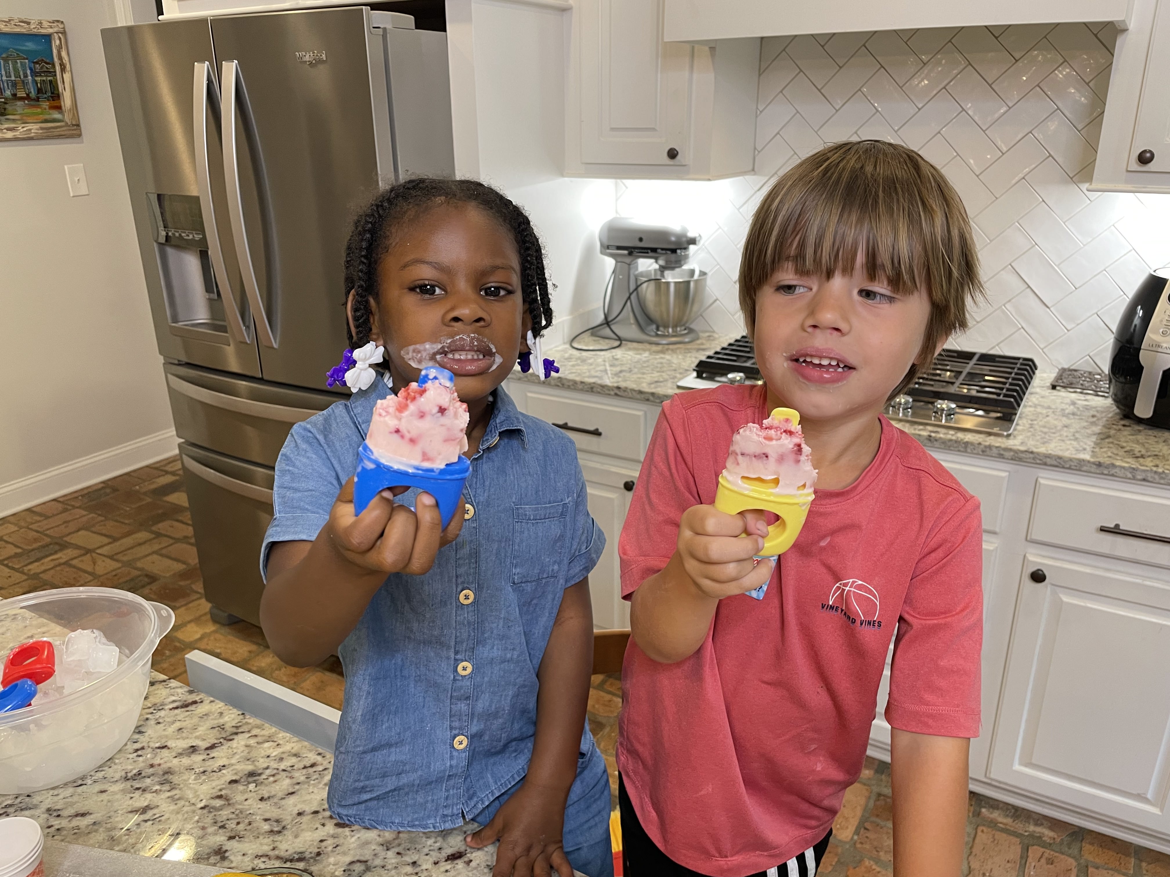 A girl and boy stand in a kitchen and hold up the popsicles they are eating.