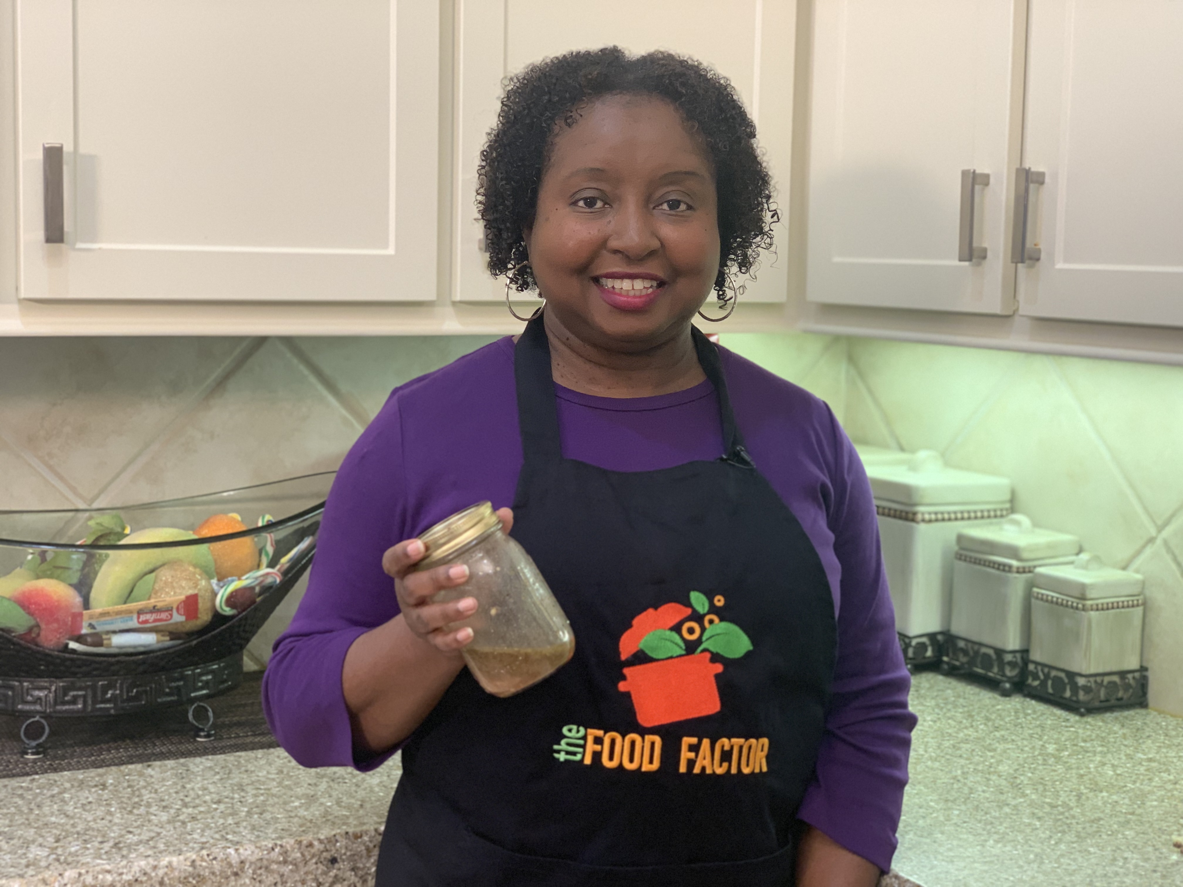 A woman stands in a kitchen and holds a jar of red wine vinaigrette.