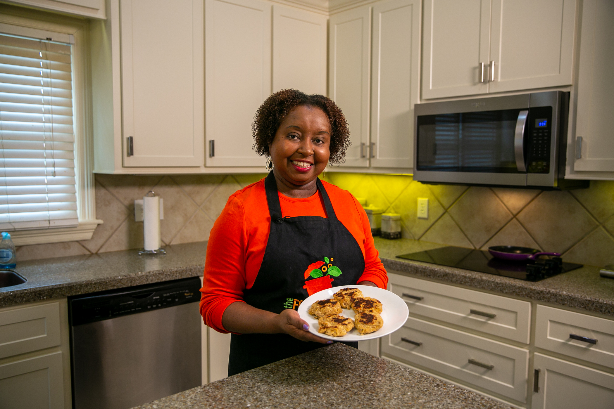A woman stands in a kitchen holding a plate of salmon patties for the camera.