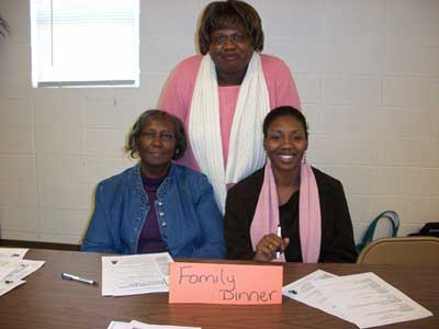 Two women sitting down and one woman standing behind them post for a picture as they gather around a small orange sign that says, "Family Dinner."