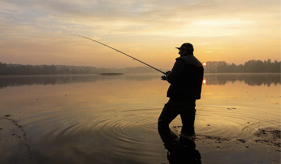 Silhouette of a man holding a casted fishing pole while standing in pondwater at sunset.