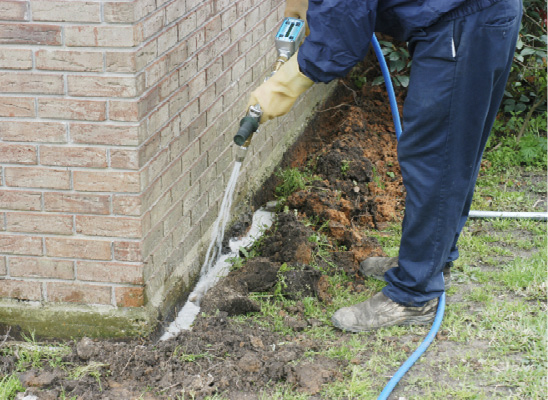 A person uses a hose-type tool to spray termite treatment into a trench next to a brick building.