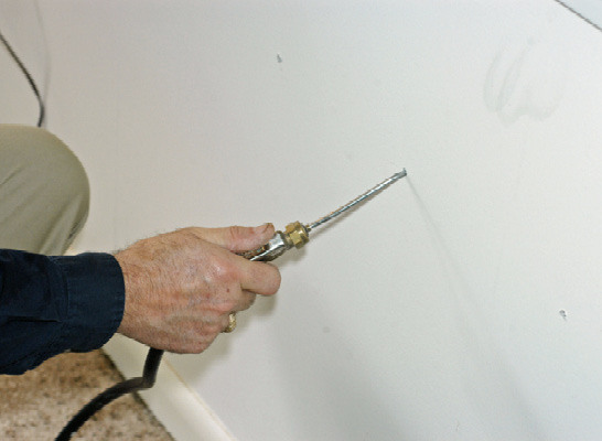 A person holds a tool with a long, narrow nozzle inserted in a tiny hole in drywall.