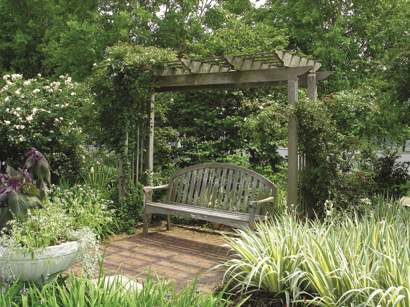 A wooden bench under an arbor sits in a brick-paved area of a garden surrounded by green grass, flowers, and trees.