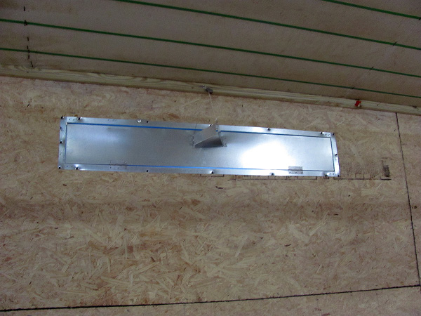 A metal plate secured to the broiler house wall with a metal flap open.