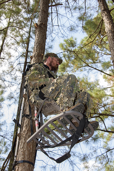 A man climbing a tree stand, off of the ground. One hand is on the stand while the other hand is on the safety strap. 