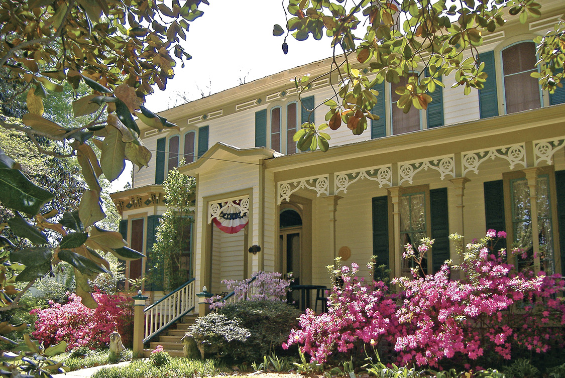 Victorian house with bright-pink azaleas and other landscaping along the front porch.