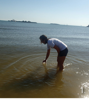 A person stands in shallow water collecting water in a jar.