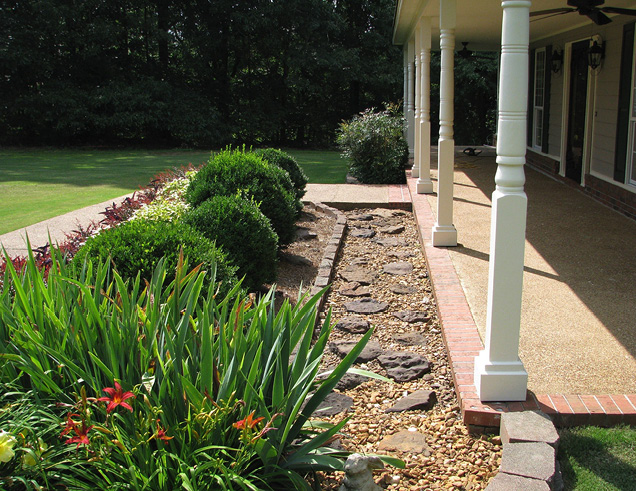 View from beside a covered front porch showing a border area filled with ornamental gravel and stones and edged with pavers. This border separates the porch from a traditional ornamental bed with shrubs, flowering plants, and ornamental grasses.