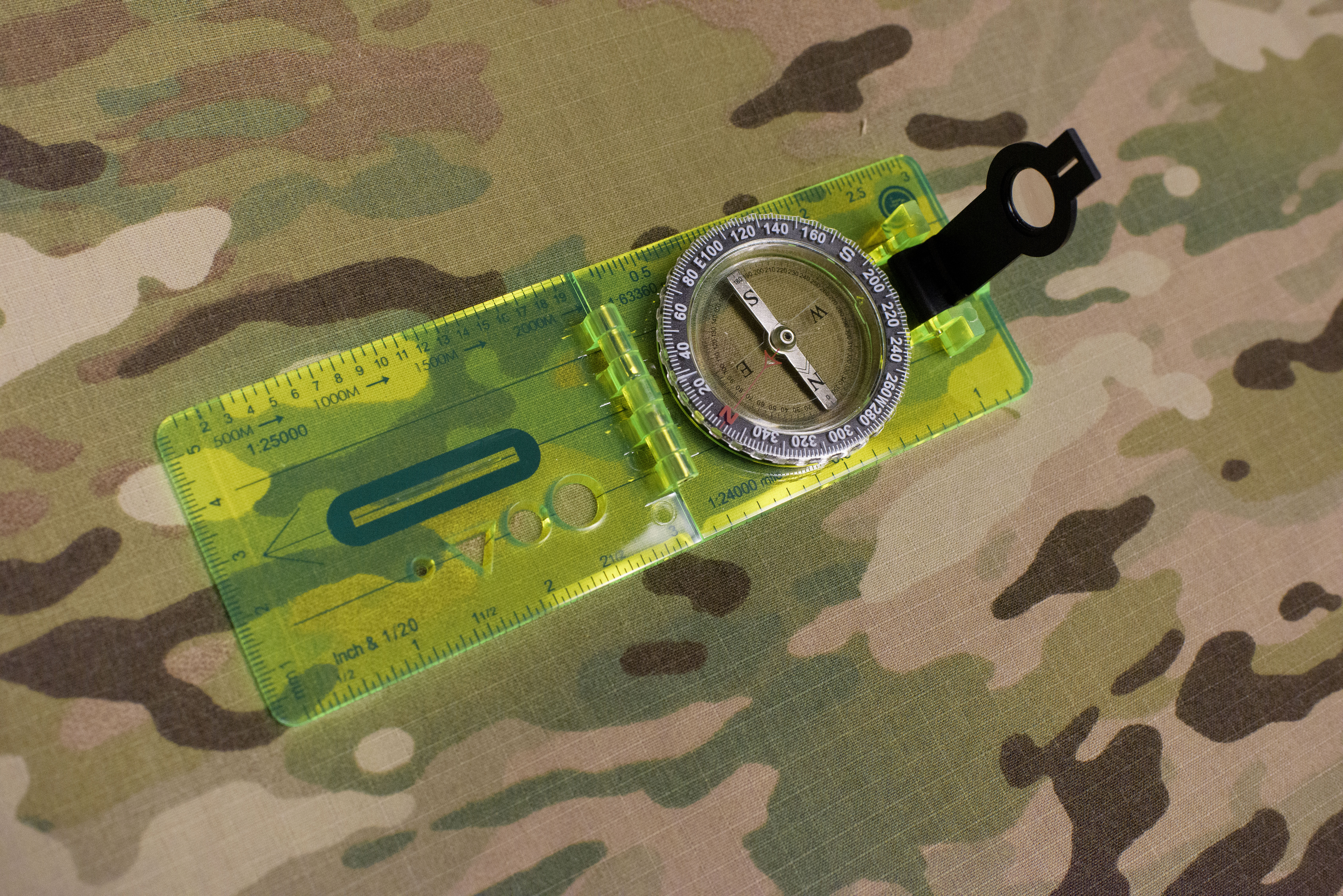 Clear rectangular ruler with an attached compass face and magnifying glass on a camouflage background.