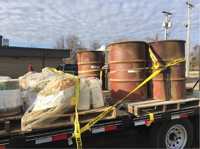 A trailer with three large, metal barrels and several plastic-wrapped containers, all secured to the trailer with ratchet straps.