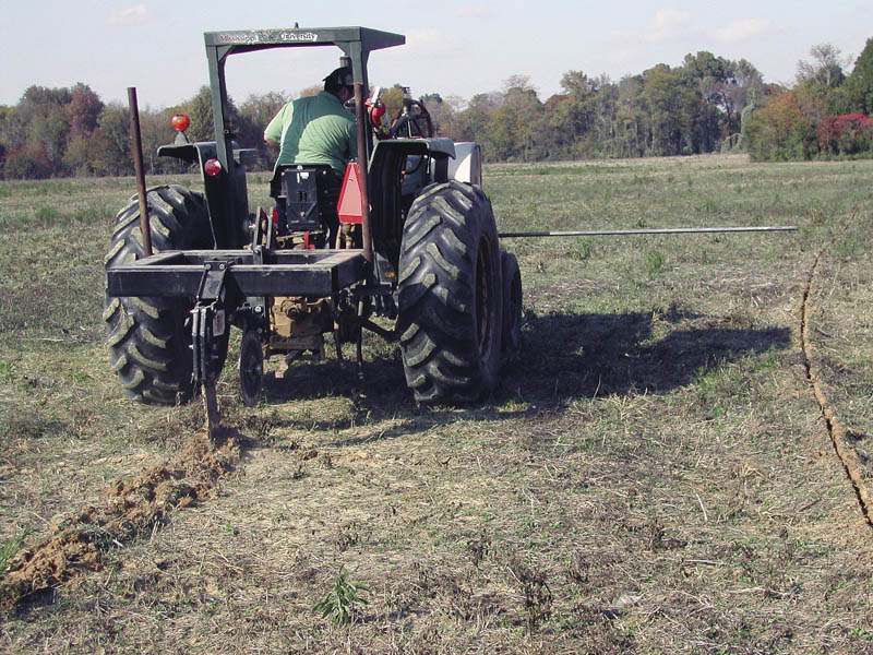 A tractor moves though a field with an attached piece of equipment that is being used to to dig a narrow trench in the soil.