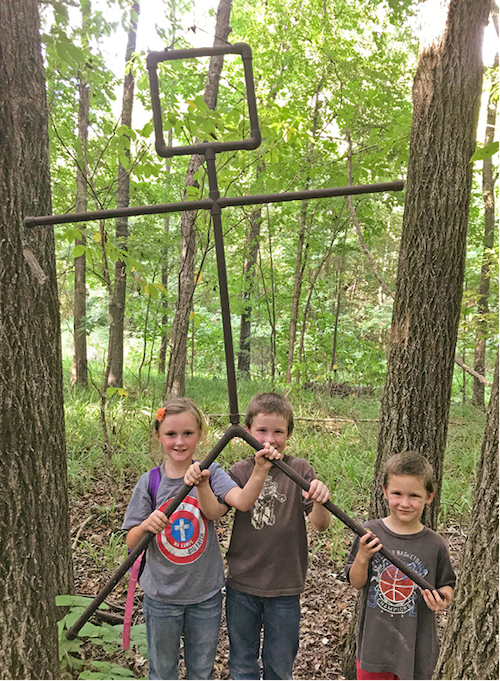 Three young children holding a large metal object that resembles a stick-figure.