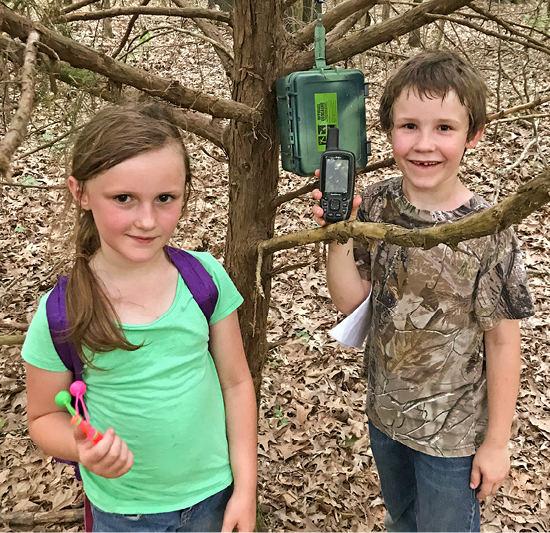 A young girl and boy holding a GPS device stand in front of a geocache hanging from a tree branch.
