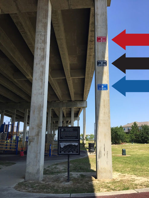A highway overpass support beam with three signs marking the high water levels from Hurricanes Hurricanes Camille and Katrina, and the base flood elevation.