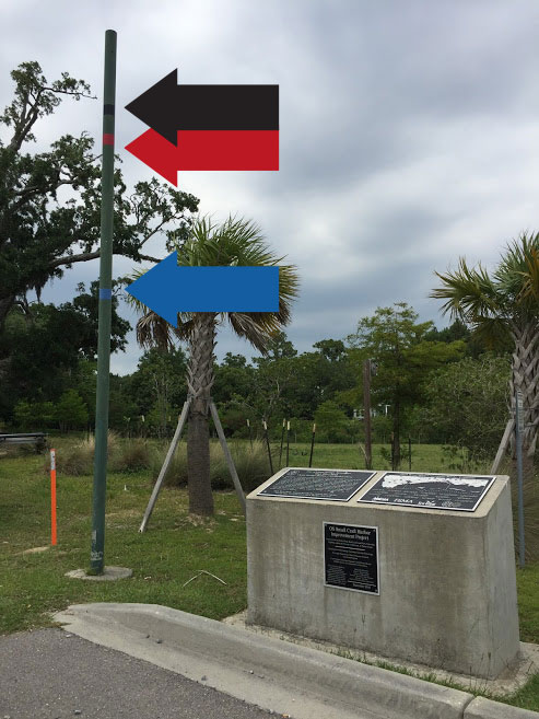 A pole with lines marking the high water levels from Hurricanes Camille and Katrina and the base flood elevation. A large concrete marker with plaques is beside the pole.