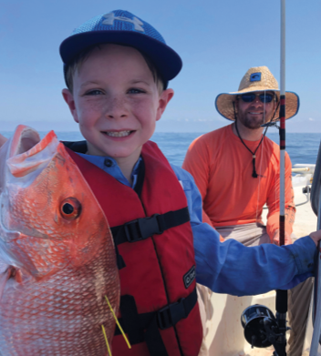 A young boy smiles at the camera as he poses on a boat in the ocean. He is holding a large orange fish with two yellow tags on its back. A man smiles in the background.
