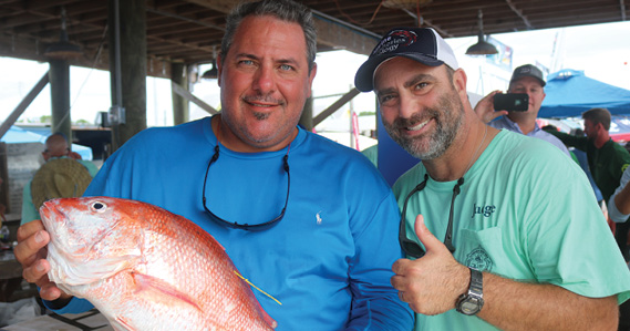 Two smiling men stand in a pavilion. One holds a large orange and white fish with a yellow tag on its back, and the other gives a thumbs-up sign.
