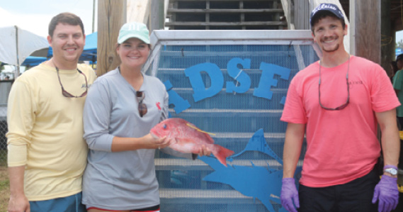 Three smiling people stand at the bottom of a staircase. One is holding a large orange and white fish with two yellow tags in its back.