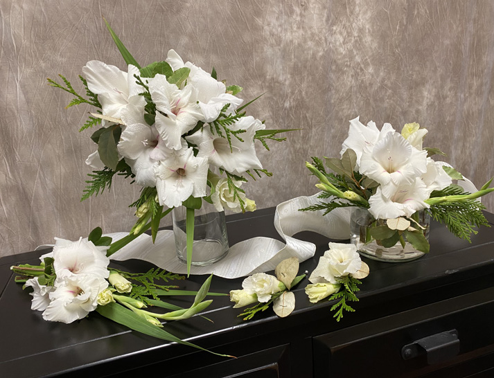 Two floral arrangements in vases, a small arrangement, and two boutonnieres displayed on a dark cabinet. The white flowers are accented with greenery. 